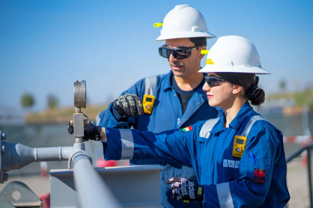 Two workers in blue coveralls and white hard hats inspecting site pipework