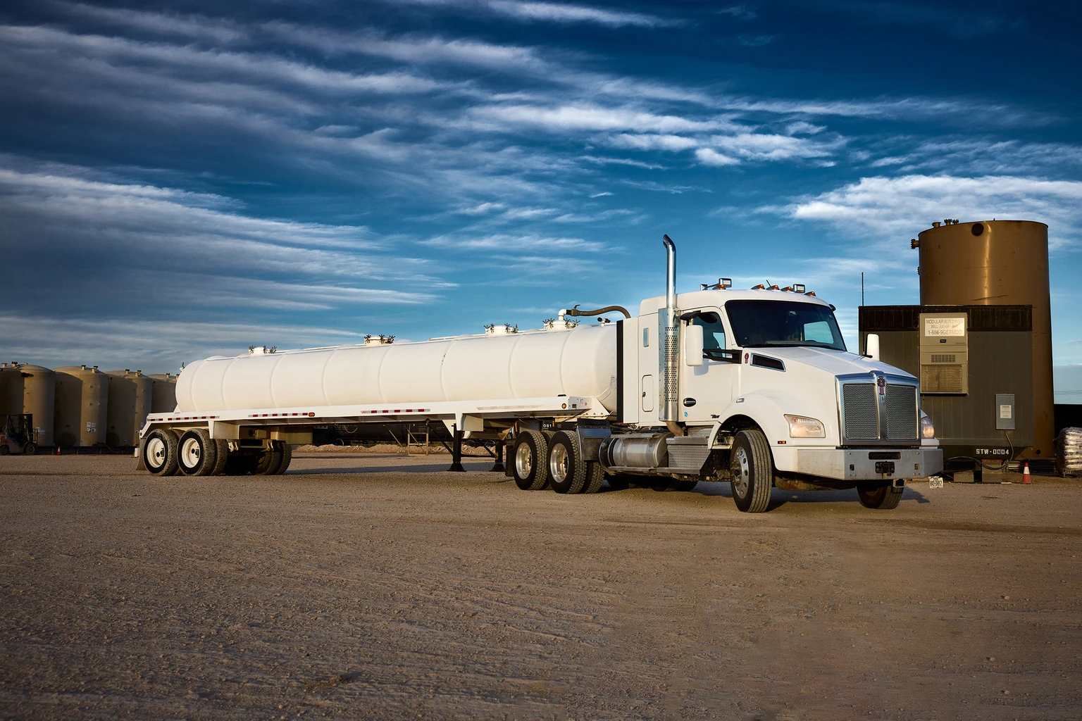 White tractor and bulk tanker parked beside industrial storage tanks